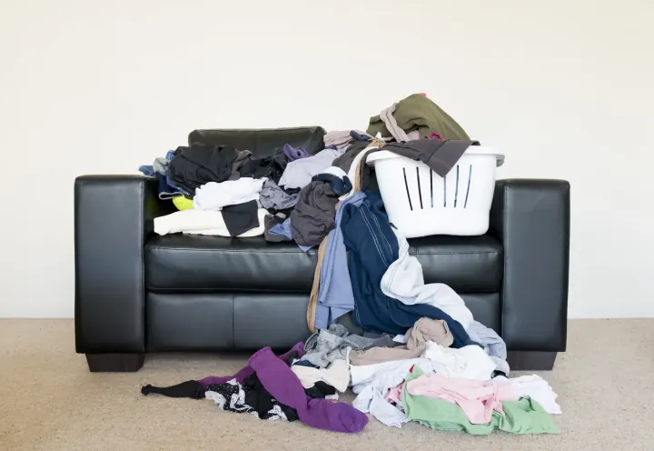 Schwarzes Ledersofa mit großem Wäscheberg aus bunten Kleidungsstücken und Wäschekorb — typisches Beispiel für beginnende Vermüllung einer Wohnung